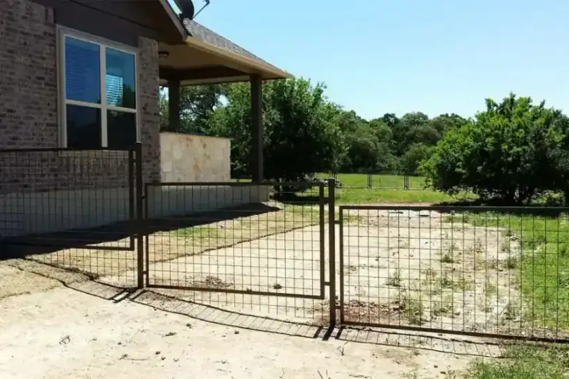 A brown welded-wire ranch fence and matching gate in front of a Hill Country house
