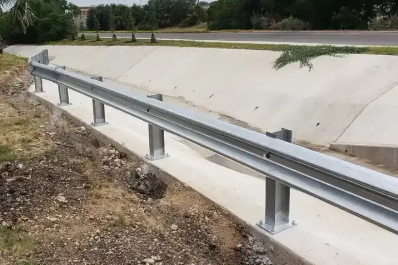 A galvanized W-beam guardrail along a concrete drainage channel, palms behind it