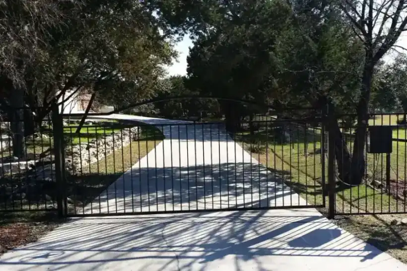 A black arched driveway gate between limestone pillars, live oak trees behind it